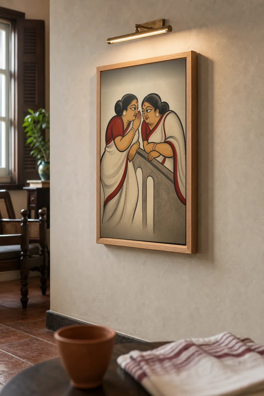 The Gossiping Kakimas Kalighat Print — two aunties trading neighbourhood gossip over a balcony rail, contemporary Bengal patua satire, hand-numbered collector edition — the framed print shown at a three-quarter angle on a styled gallery wall in a natural-oak frame