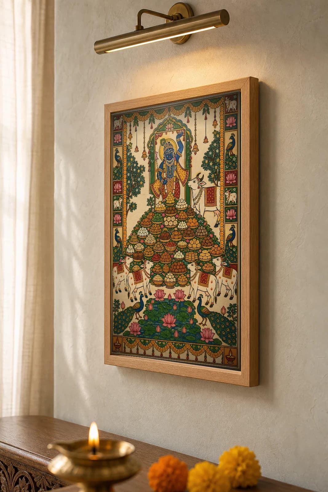 Annakut Feast Mountain Pichwai-Style Print — Shrinathji above a pyramid of food bowls with cows, peacocks and a lotus pond, on warm cream cloth — the framed print shown at a three-quarter angle on a styled gallery wall in a natural-oak frame