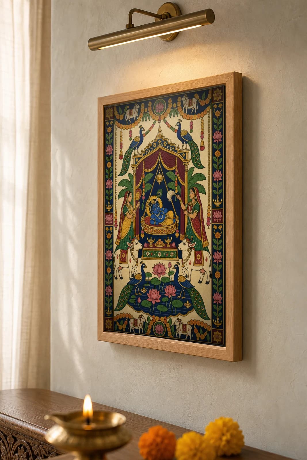 Janmashtami Jhanki Pichwai-Style Print — baby Krishna in an ornate swing-cradle under a canopy, attended by two women, with cows, peacocks and a lotus pond, on warm cream cloth — the framed print shown at a three-quarter angle on a styled gallery wall in a natural-oak frame